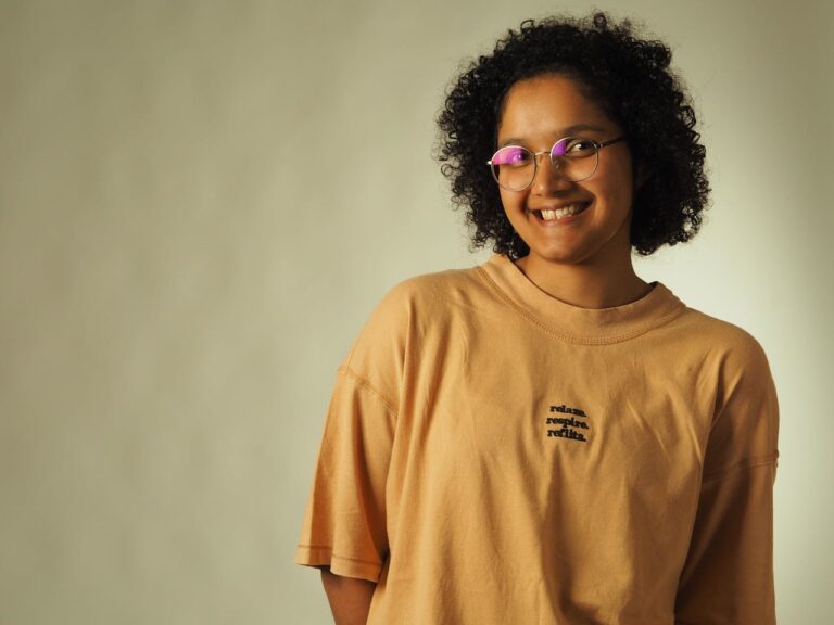 Cheerful woman with curly hair and glasses wearing a beige t-shirt with text.